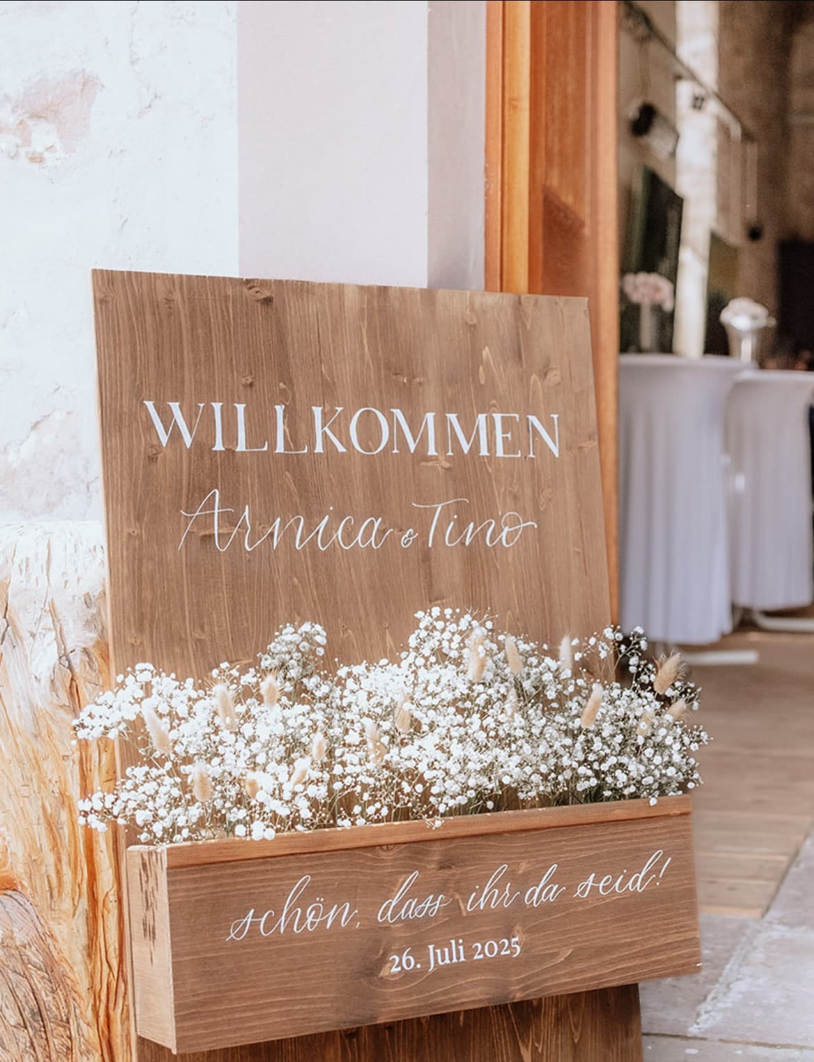 Hölzernes Willkommensschild mit weißer Kalligrafie, Namen und Hochzeitsdatum, dekoriert mit Trockenblumen