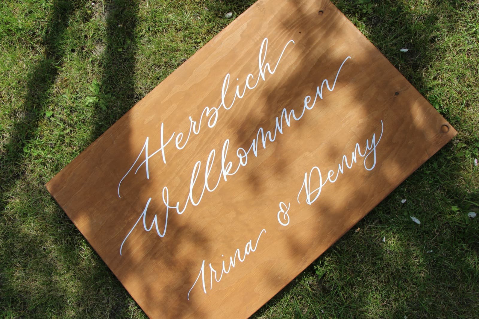 Hölzernes Willkommensschild mit der Aufschrift Herzlich Willkommen Trina und Denny in weißer Kalligrafie auf einer Wiese